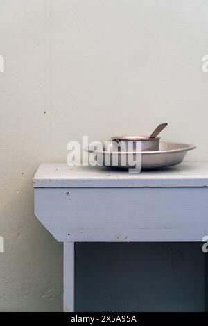 A stainless steel frying pan with a spoon rests on a simple white wooden table against a plain beige wall, evoking a sense of simplicity and minimalis Stock Photo