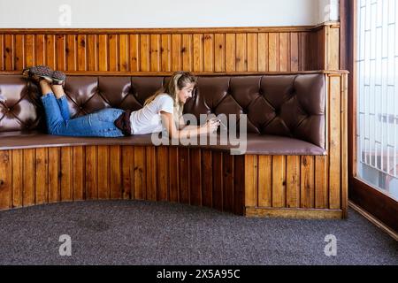 Young woman relaxing and using smartphone on a vintage leather bench with wood paneling interior. Stock Photo