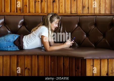 A young woman lies on a bench, deeply engrossed in her smartphone, in a room with wooden walls. Stock Photo