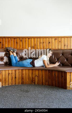 A young woman lies on her side on a classic diner-style bench, engrossed in her smartphone, with a wood-paneled wall behind her. Stock Photo