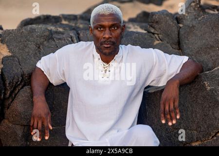 An elegant African American senior man poses on a beach, exuding tranquility against a backdrop of rocks and sand Stock Photo