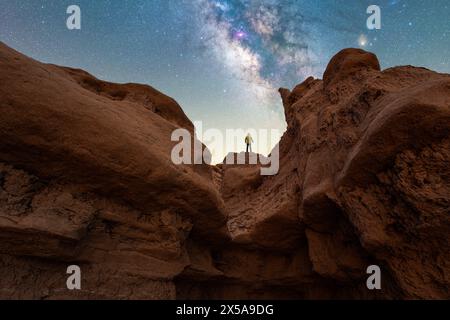 An adventurer stands atop a formation in Goblin Valley State Park, Utah, with the Milky Way shimmering above in the night sky. Stock Photo