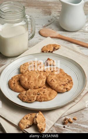 A cozy arrangement of chocolate chip cookies on a ceramic plate, paired with a glass jar of milk and a wooden spoon, all portrayed on a rustic wooden Stock Photo