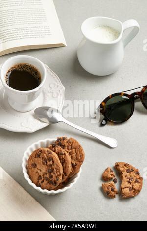 A cozy tabletop view featuring a cup of coffee, cookies on a plate, and a milk jug, accompanied by reading glasses and an open book Stock Photo