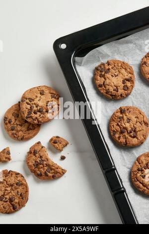 Freshly baked chocolate chip cookies cooling on a black baking tray lined with parchment paper, some broken, showcasing a tempting treat Stock Photo