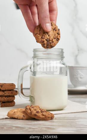A hand captured mid-motion as it prepares to dunk a chocolate chip cookie into a jar of fresh milk, with more cookies and a white backdrop Stock Photo