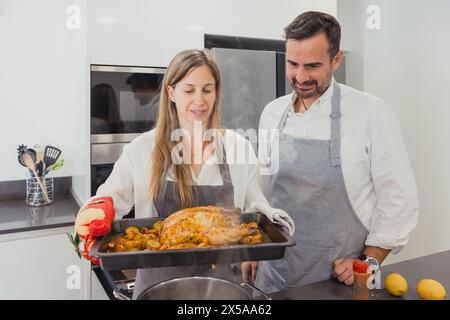 A smiling man and woman wearing aprons stand in a modern kitchen, taking a roasted chicken with vegetables out of the oven Stock Photo