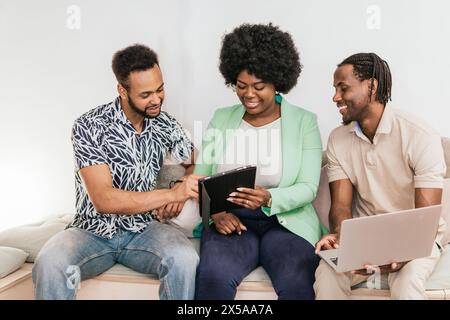 Three young professionals share ideas using a tablet and a laptop while seated on a couch in an informal coworking space Stock Photo