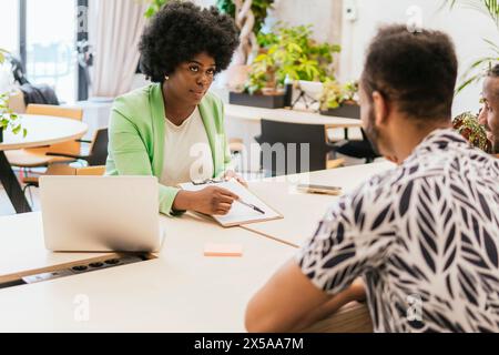 Two professionals engage in a discussion at a sunlit coworking space, with a woman taking notes and a laptop open Stock Photo