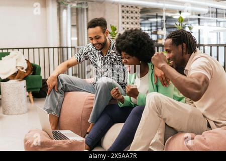 Three colleagues cheer in excitement in a modern coworking space while looking at the laptop screen Stock Photo