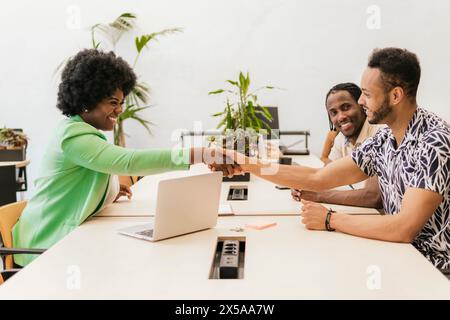Three professionals happily engaging in a fist bump over a work table in a bright coworking environment Stock Photo