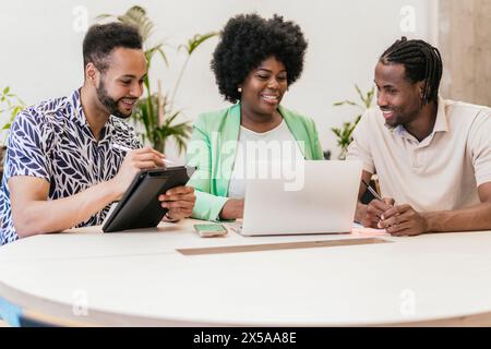 Three professionals engage in a collaborative meeting using a laptop and tablet in a coworking space featuring modern, bright decor Stock Photo