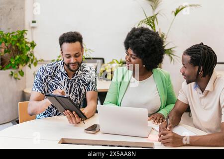 Three professionals collaborate at a coworking space, sharing ideas on a digital tablet and laptop Stock Photo