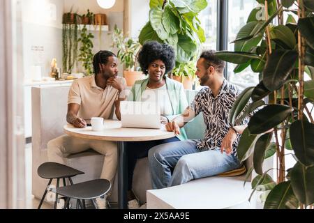 Three colleagues engage in discussion around a laptop in a bright and plant-filled coworking environment Stock Photo