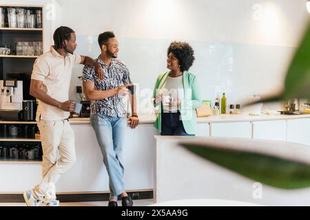 Three colleagues converse and drink coffee during a casual break in a well-lit, contemporary office kitchen, showcasing camaraderie and a collaborativ Stock Photo