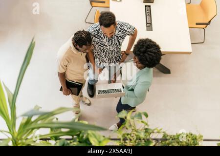A top-view image of three professionals standing in a coworking office, engaging in a discussion with a laptop Stock Photo
