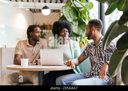 Three professionals collaborating around a laptop in a plant-filled, modern co-working environment Stock Photo