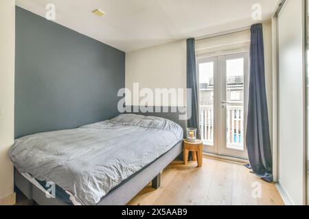 A modern minimalist bedroom featuring a large bed with grey bedding, wooden floor, and natural light from a window with blue curtains. Stock Photo