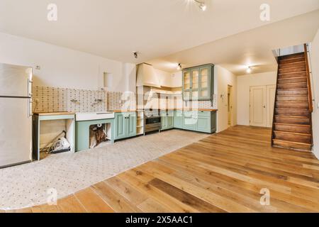 A wide-angle view of a spacious kitchen with mint-colored cabinets, modern appliances, a wooden staircase, and hardwood flooring. Stock Photo