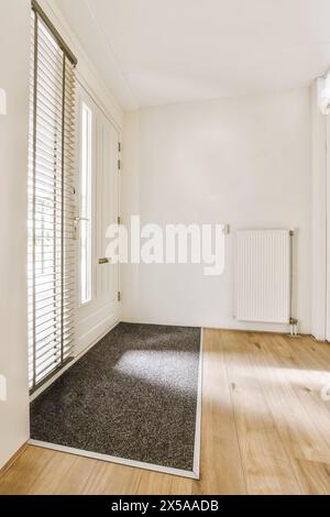 A bright, empty entryway featuring white walls, wooden floor, a radiator, and venetian blinds casting shadows. Stock Photo