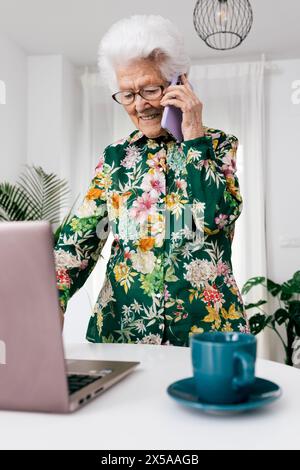An elderly woman in a floral blouse is working from home, using a laptop and conversing on a phone Stock Photo