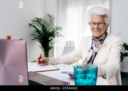 A cheerful elderly woman engaging in work from her home office, holding a pen and writing on notepad with a laptop open in front of her Stock Photo