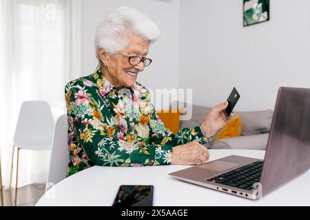 Cheerful elderly woman in a floral shirt joyfully using a credit card to shop online on her laptop at home Stock Photo