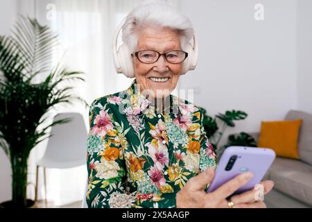 A cheerful elderly woman with white hair is wearing large headphones and using a smartphone at home, her smile exuding happiness Stock Photo