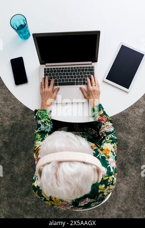An elderly woman in a floral shirt is seen from above working on her laptop at home, with phone and tablet nearby Stock Photo