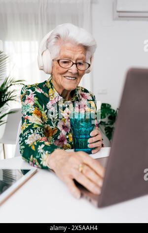 Elderly lady in a floral shirt smiles while using a laptop and wearing headphones, exemplifying active aging and technology use at home Stock Photo