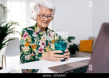 Elderly lady in a floral shirt enjoys a glass of water while working on her laptop in a bright living room, showcasing active aging and technology use Stock Photo