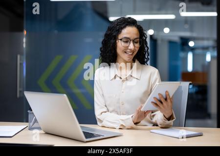 Young successful businesswoman at workplace is satisfied with results of achievement at work smiling and using tablet computer, programmer is testing new software in application Stock Photo