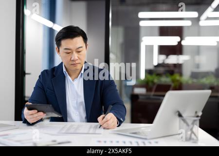 Focused Asian businessman reviewing financial reports while working in a modern office space. Concept of professionalism and diligence. Stock Photo