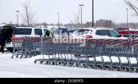 A row of grocery shopping carts on a winter day in a snow covered parking lot with many vehicles. Stock Photo