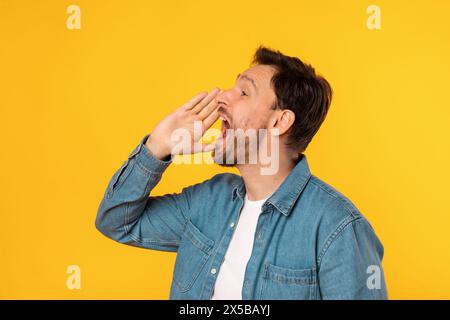 Young Man in Denim Shirt Shouting Announcement Against Yellow Background Stock Photo
