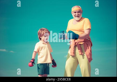 Portrait of a determined senior boxer with little boy child over blue sky background. Boxer grandfather and child with blue boxing glove on blue sky Stock Photo