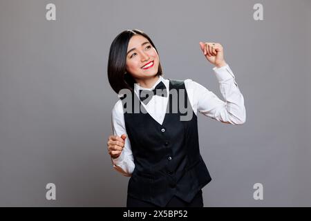 Attractive carefree asian woman receptionist wearing restaurant uniform joyfully dancing and having fun. Cheerful smiling young waitress moving arms to dynamic music beats Stock Photo