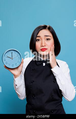 Upset asian waitress showing alarm clock and looking at camera with disappointed facial expression. Sad woman receptionist checking time while running late studio portrait on blue background Stock Photo