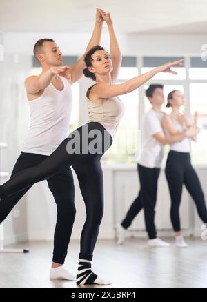Pretty female ballerina and male partner practicing ballet arabesque pose during training with group active people in dancing hall Stock Photo