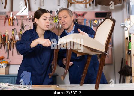 Senior male furniture maker and female apprentice working on vintage commode in workshop Stock Photo
