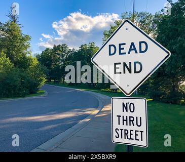 White Warning Road Signs on DEAD END street and NO THRU STREET along road and sidewalk in evening. Stock Photo