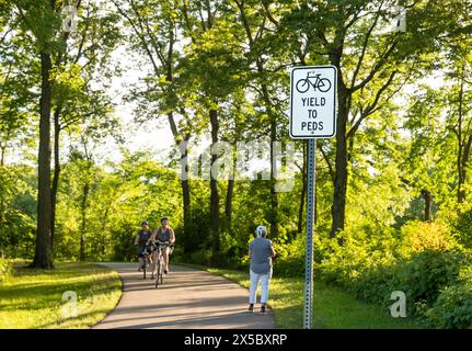 YIELD TO PEDS Sign on the bike and walking path in a wooded area with paved trail and green trees, and a pedestrian meeting a couple bikers. Stock Photo