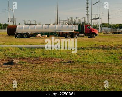 Wide view over green grass to a steel Utility Pole section laying ground a with a water truck and Electric Substation in back. afternoon. Blue sky. Stock Photo