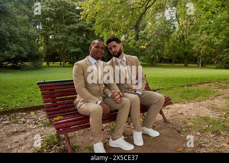 Gay newlywed couple sitting on a wooden bench holding hands in a natural environment. They are wearing formal suits, newlywed couple. Stock Photo