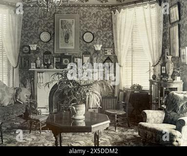 A front room or parlour of a British house c.1900. The admittance of direct sunlight is kept under control by the adjustable slatted Venetian blinds at the windows, which also have net curtains hung from a pole. The room is full of ornaments and the table has an indoor plant on it. Although often viewed as being ‘modern’, Venetian Blinds date back to mid-1700s Venice. Their popularity initially spread to France – the French refer to the blinds as les Persiennes. Early Venetians were made of wood slats and suspended on vertical cloth ribbons. They provided an alternative or addition to curtains Stock Photo