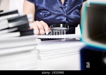 An office worker examines documents through a magnifying glass against the background of stacks of documents Stock Photo