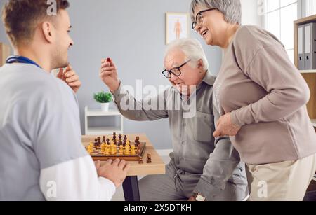 Young nurse man playing chess game with happy smiling senior elderly couple in clinic. Stock Photo