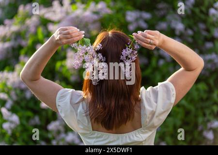 A girl with loose hair holds lilac flowers in her hands and stands with her back to the camera. Stock Photo