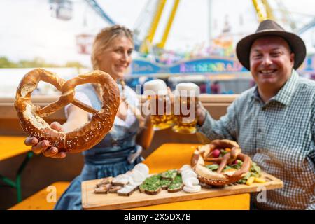Woman holding a pretzel and toasting beer mugs with a man at a beer tent table with snacks on a wooden board and amusement rides at oktoberfest or dul Stock Photo
