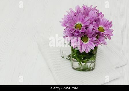 A clear glass vase filled with water sits on a white table. The vase holds a bouquet of purple chrysanthemum flowers with green stems and leaves. Stock Photo